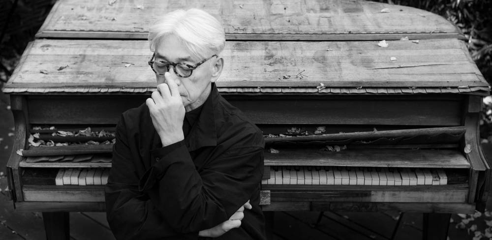 Ryuichi Sakamoto, Portrait – Schwarzweiß-Fotografie eines nachdenklichen Menschen mit Brille und hellem Haar vor einem alten, teils kaputten Klavier mit Herbstblättern.