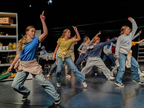 Zum Lachen in den Keller, LABYRINTH Theater Stuttgart – Auf dem Bühnenfoto sind etwa zehn weiblich gelesene Jugendliche abgebildet. Sie tragen Jeans und lockere, bunte Oberteile. Sie wirken, als würden sie gerade tanzen oder sich dynamisch bewegen, da einige Füße leicht in der Luft sind und die Arme ausgestreckt nach oben rechts zeigen. Ihre Körper sind nach vorne gerichtet, sie schauen jedoch ebenfalls nach rechts. Im Hintergrund sind Regale mit Dosen und Kisten sowie eine Leiter zu sehen.