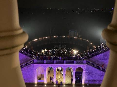 Adventsstimmung – Blick durch Balustrade auf beleuchteten Innenhof, mit Menschenansammlung vor Arkaden und Weihnachtsbaum; Nachtaufnahme.