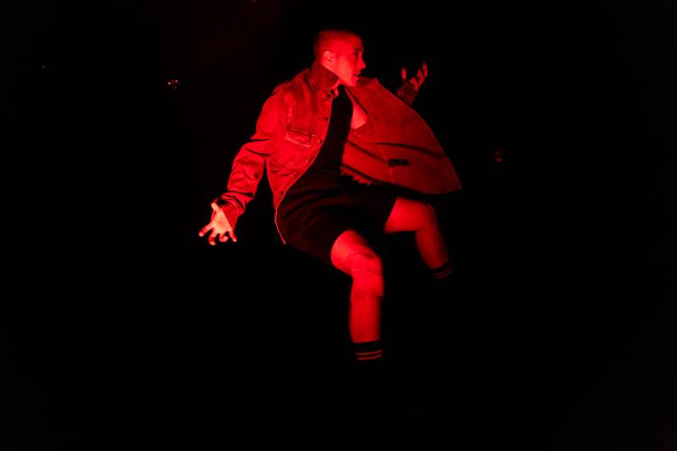 Eine Person tanzt in rotem Bühnenlicht vor dunklem Hintergrund.