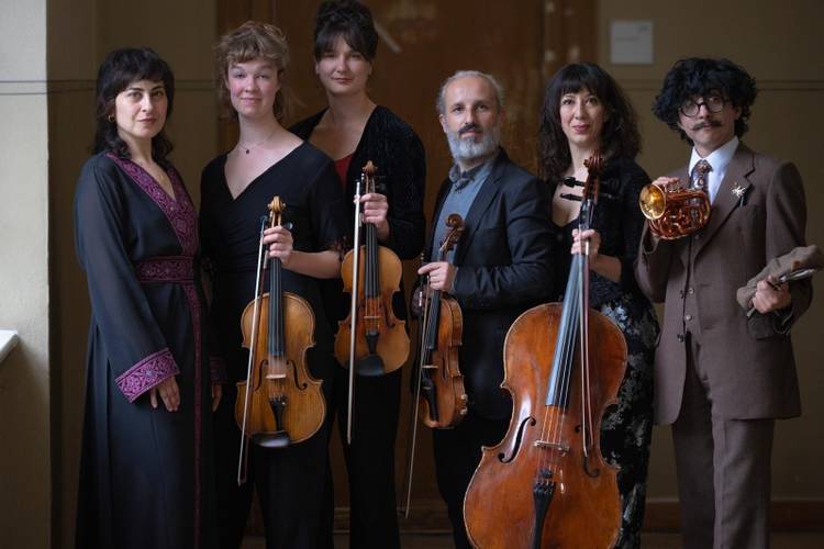 Sechs Personen in eleganter Kleidung mit Instrumenten: drei Violinen, ein Cello, ein Horn. Person rechts hält ein Buch. Sie stehen in einem Raum.