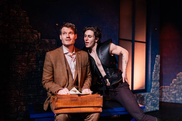Dracula – A Comedy of Terrors – Zwei Personen auf der Bühne. Eine trägt Anzug und hält einen Kasten, die andere trägt eine offene Rüstung. Beide interagieren miteinander.