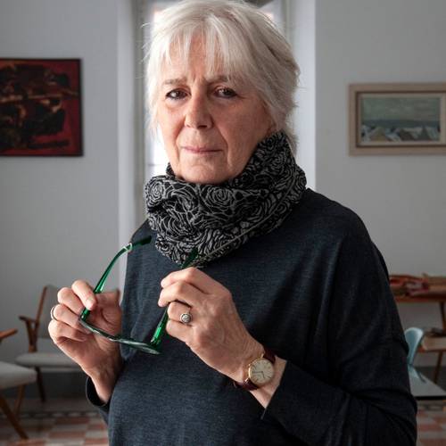 Anita Raja – Ältere Person mit grauem Haar hält eine Brille in der Hand, trägt einen gemusterten Schal und eine Uhr. Im Hintergrund sind Stühle und Bilder zu sehen.
