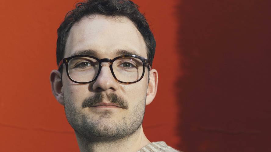 Lukas Rietzschel – Person mit Brille und Bart vor rotem Hintergrund, schaut direkt in die Kamera. Person hat kurzes dunkles Haar.