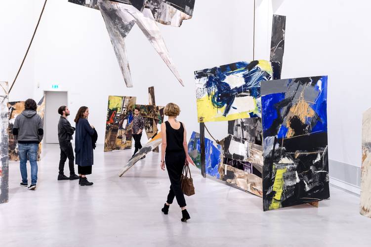 Installationsansicht „Emilio Vedova. Absurdes Berliner Tagebuch ’64“, 1964, Berlinische Galerie © Foto: Lutz Bertram, © Fondazione Emilio e Annabianca Vedova – Foto von Menschen im Ausstellungsraum zwischen Teilen der Installation. Dabei stehen großformatige, abstrakte Installationsobjekte aus bemalten Holzplatten im Ausstellungsraum. Die Platten sind mit kräftigen Pinselstrichen in Schwarz, Weiß, Grau, Gelb und intensivem Blau gestaltet und in verschiedenen Winkeln im Raum positioniert. Der Boden ist glatt und hell, die Wände neutral und ohne zusätzliche Elemente.