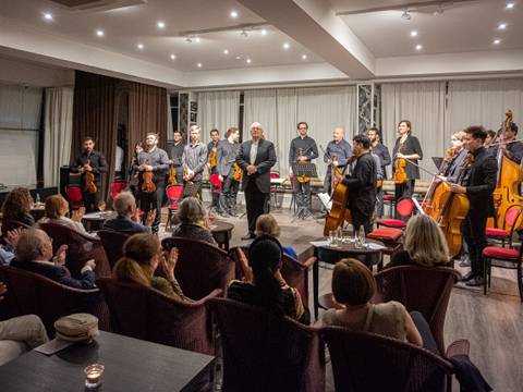  – Eine Gruppe von Musiker*innen mit Streichinstrumenten steht verneigend vor sitzendem Publikum in einem Raum mit roten Stühlen und hellen Vorhängen.