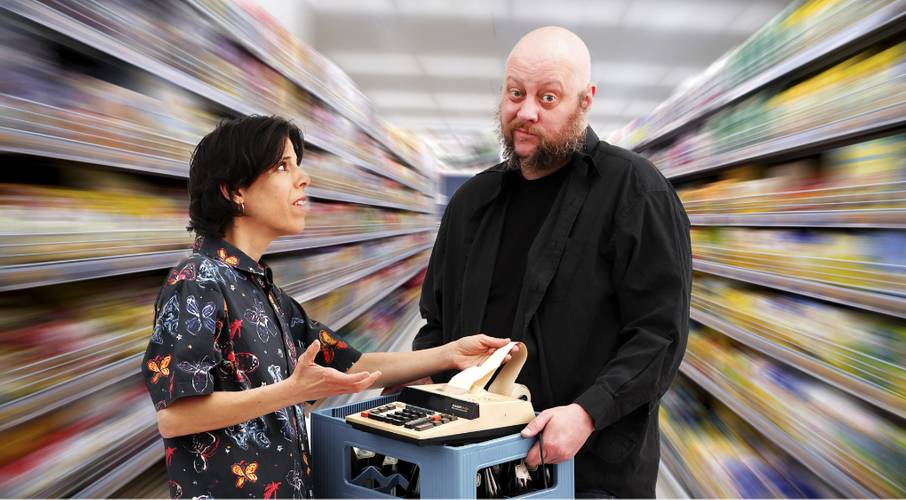 Eine Person mit Hemd mit Schmetterlingsmotiv spricht in einem Supermarkt mit einer männlich gelesenen Person, die ein Kassengerät hält.