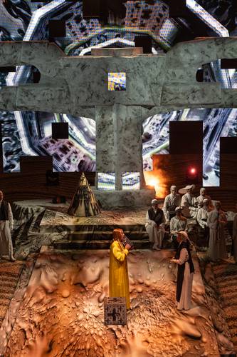 Mittwoch aus Licht – Bühne mit gemustertem Boden; zehn Personen in mittelalterlicher Kleidung; zentrale Person im gelben Kleid hält Buch; steinerner Hintergrund mit Projektionen.