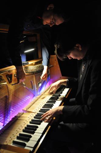 SOHLE EINS-ACHTZIG – Zwei Personen, eine spielt Klavier, die andere schlägt zwei Löffel im Inneren auf die Saiten. Kunstvolle violette Beleuchtung im Klavier.