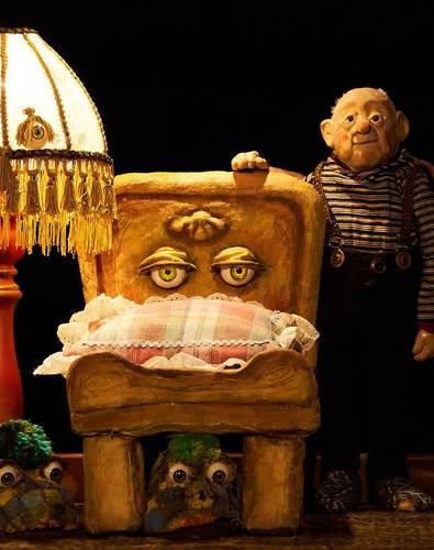 Männlich gelesene Figur in gestreiftem Hemd neben einem Stuhl mit Gesicht, Lampe links, Puppen unter dem Stuhl.