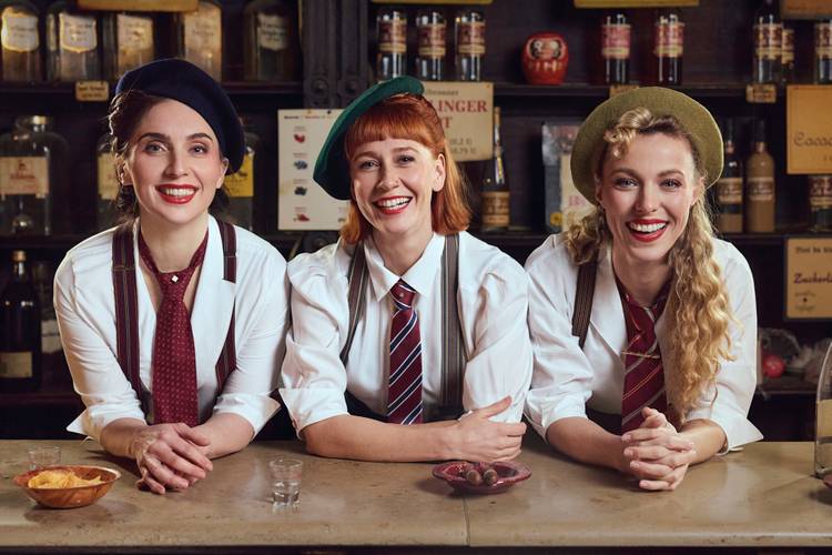 The Airlettes: Live – Drei lachende Personen in Hemden, Krawatten und Trägern hinter Tresen. Zwei tragen Baskenmützen, eine trägt einen hellgrünen Hut. Tisch hat Schale und Gläser.