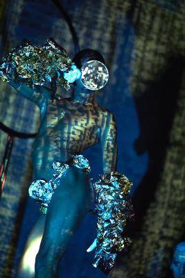 Queer Performance Festival – Person mit verspiegeltem Gesicht und metallischer Dekoration an Armen und Hüfte, beleuchtet von bunten Mustern. Hintergrund dunkel mit Schatten.