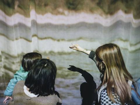 Foto: Nancy Jesse – Eine Familie steht vor einem Vorhand auf den Streifenmuster projiziert sind. Ein Kind macht hält seine Hand so, dass der Schatten einen Tierumriss auf den Vorhang wirft.