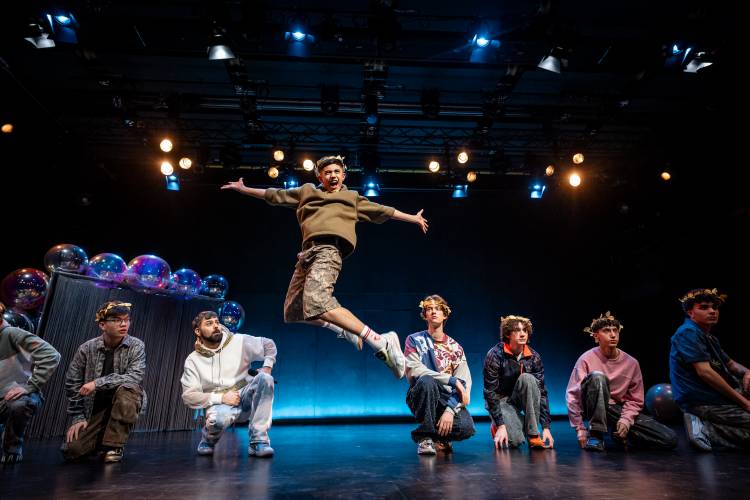 APOLLON - STOP TRYNA BE GOD, waltraud900, Forum Freies Theater Düsseldorf – Ein Bühnenfoto in dem acht männlich gelesene Personen abgebildet sind. Sieben von ihnen knien auf dem Boden und schauen größtenteils gerade aus. Der in der mitte springt gerade und befindet sich mit weit ofnnen Armen und einem schreienden Gesichtsausdruck in der Luft. Im Hintergrund sind Scheinwerfer wodurch eine theatrale Stimmung entsteht. Sie tragen typische jugendliche Kleidung und tragen einen goldenen Lorbeerkranz auf dem Kopf.