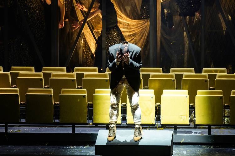 Rigoletto – Person in glitzernder Kleidung beugt sich nach vorne, haelt den Kopf in den Haenden, umgeben von gelben Sitzen, dahinter spiegelbildliche Silhouetten.