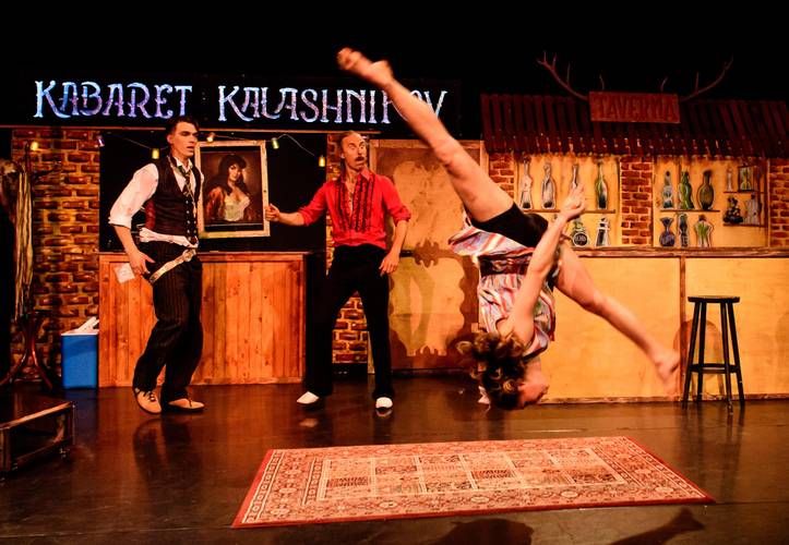 Kabaret Kalashnikov – Person macht Handstand in bunter Kleidung; zwei Personen in Kostümen schauen zu; Hintergrund mit "KABARET KALASHNIKOV" Schild und Bar-Dekor.