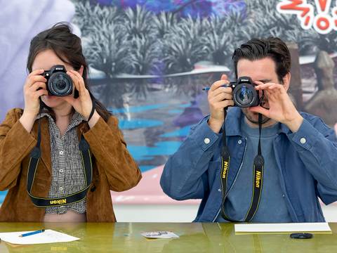  – Zwei Personen fotografieren mit Nikon Kameras. Im Hintergrund ein farbenfrohes Gemälde mit Ausrufezeichen. Auf dem Tisch liegen Papier und Stifte.