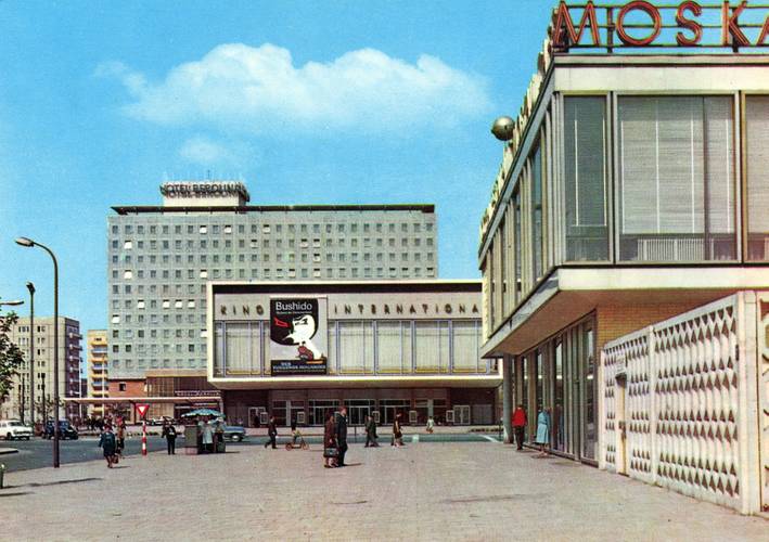 Postkarte, Karl-Marx-Allee Berlin mit dem Ensemble Kino International, entworfen von Josef Kaiser und Heinz Aust, Hotel Berolina, entworfen von Josef Kaiser und Günter Kunert, sowie Restaurant Moskau, entworfen von Josef Kaiser und Horst Bauer, 1964 – Straßenszene mit mehreren Gebäuden: Im Hintergrund steht ein hohes, rechteckiges Hotelgebäude mit vielen gleichförmigen Fenstern und einem Schild „Hotel Berolina“ auf dem Dach. Davor befindet sich ein breiter, niedriger Bau mit großen Fensterfronten und einer Filmwerbung an der Fassade. Rechts im Bild ist ein modernes Gebäude mit großen Glasfronten und einem Schriftzug auf dem Dach zu sehen. Auf dem Platz davor gehen verschiedene Personen, einige Autos stehen oder fahren im Hintergrund. Der Himmel ist klar und blau.