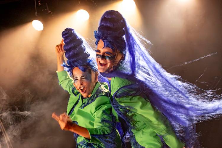 oh peinlich – Zwei Personen mit blauen Perücken und grünen Kostümen, tanzend auf beleuchteter Bühne. Rauch umgibt sie, beide lachen und gestikulieren lebhaft.