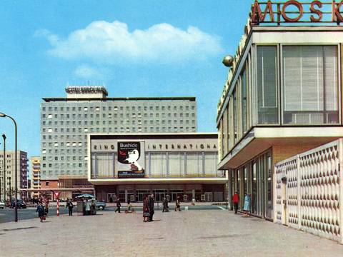 Postkarte, Karl-Marx-Allee Berlin mit dem Ensemble Kino International, entworfen von Josef Kaiser und Heinz Aust, Hotel Berolina, entworfen von Josef Kaiser und Günter Kunert, sowie Restaurant Moskau, entworfen von Josef Kaiser und Horst Bauer, 1964 – Straßenszene mit mehreren Gebäuden: Im Hintergrund steht ein hohes, rechteckiges Hotelgebäude mit vielen gleichförmigen Fenstern und einem Schild „Hotel Berolina“ auf dem Dach. Davor befindet sich ein breiter, niedriger Bau mit großen Fensterfronten und einer Filmwerbung an der Fassade. Rechts im Bild ist ein modernes Gebäude mit großen Glasfronten und einem Schriftzug auf dem Dach zu sehen. Auf dem Platz davor gehen verschiedene Personen, einige Autos stehen oder fahren im Hintergrund. Der Himmel ist klar und blau.