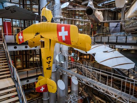 Die Bücker Bü 131 ist eines der ersten und weltweit bekanntesten Kunstflugzeuge. – Ein großes gelbes Flugzeug hängt senkrecht im offenen Treppenhaus des Neubaus. Im Hintergrund hängen weitere Flugzeuge und Flugapparate.