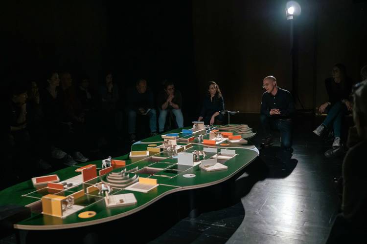The Game – Eine Gruppe von Menschen sitzt im Dunkeln um ein beleuchtetes Architekturmodell auf einem Tisch. Eine männlich gelesene Person spricht.