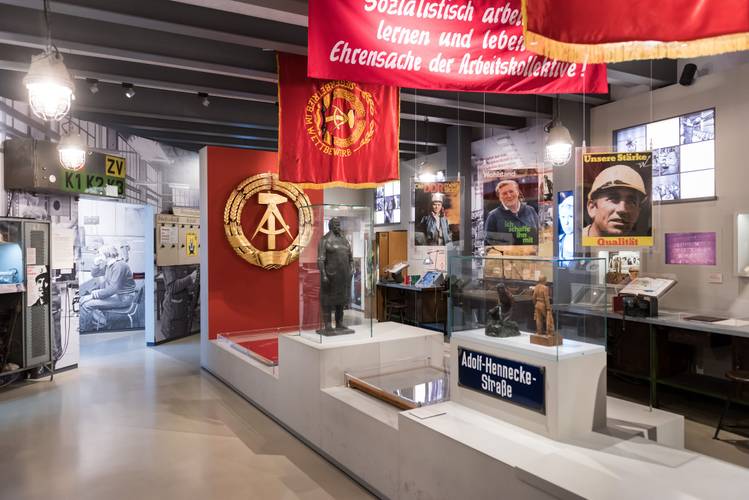Dauerausstellung "Alltag in der DDR" – Blick in einen Raum der Ausstellung "Alltag in der DDR", zwischen Vitrininen mit Exponanten hängen Transparente, Fahnen und Propagandaplakate aus der DDR