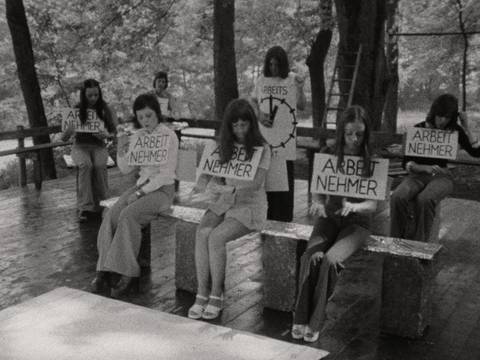 ›Helfen können wir uns nur selbst‹, BRD 1974, Regie: Gardi Deppe © Deutsche Kinemathek – Schwarz-weiß-Szenenfoto: Junge frauen sitzen auf einer Bank in der Natur, sie machen Tipp-Bewegungen mit den Händen, als würden sie Schreibmaschine schreiben. Um den Hals haben hängen Schilder auf denen das Wort Arbeitnehmer steht.
