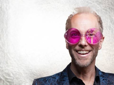 Michael Krebs: OPTIMISMUS – jetzt stark reduziert – Person mit rosa Brille und buntem Jackett vor hellem Hintergrund, lächelt in die Kamera.