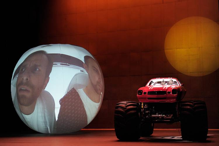 Die Entführung aus dem Serail – Menschliche Gesichter auf aufgeblasenem Globus projiziert, roter Monstertruck daneben mit zwei Personen innen, orange beleuchteter Hintergrund.