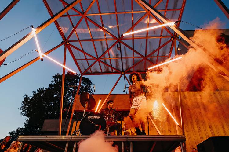 Mélissa Guex – Foto: Mélissa Guex steht auf einer überdachten Bühne unter freiem Himmel. Die Bühne wird von orangefarbenem, warmen Licht angestrahlt.
