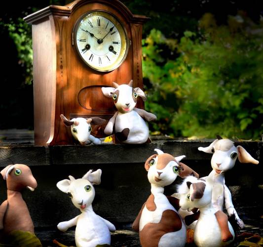 Sechs Plüschziegen vor einer antiken Standuhr mit römischen Ziffern im Freien.