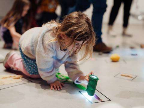  – Foto: Ein Kind sitzt mit Taschenlampe zwischen kreativem Material im Ausstellungsraum auf dem Boden.