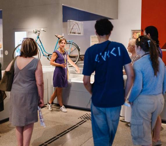 Eine Guide ist in der Mitte des Bildes zu sehen und zeigt auf eine Glasvitrine. An den Rändern des Bildes sind Teilnehmende der Führung zu sehen. Im Hintergrund sieht man ein Fahrrad und eine Säge, die zwei Objekte der Ausstellung sind.