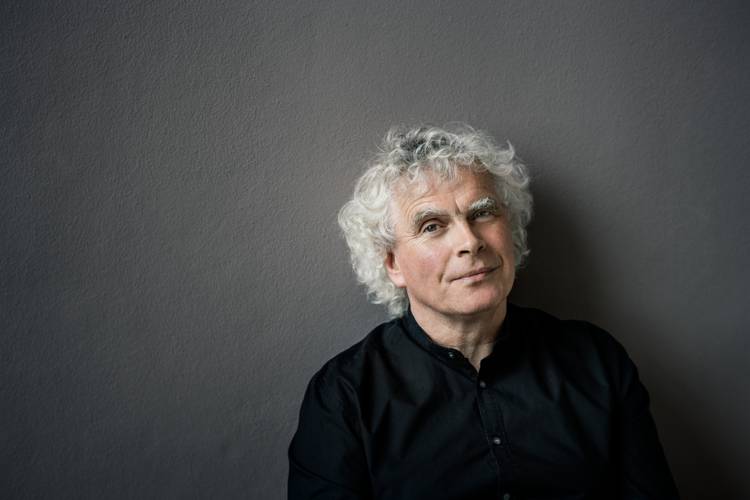 Sir Simon Rattle – Ein Mann mit weißen Locken und einem schwarzen Hemd steht vor einer neutralen Wand. Sein Blick in die Kamera ist entspannt.