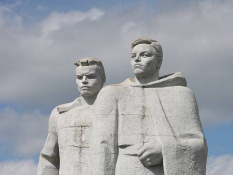  – Zwei steinerne Statuen von maennlich-gelesenen Personen, nebeneinander stehend, mit ernstem Ausdruck. Hintergrund aus Wolken und blauem Himmel.