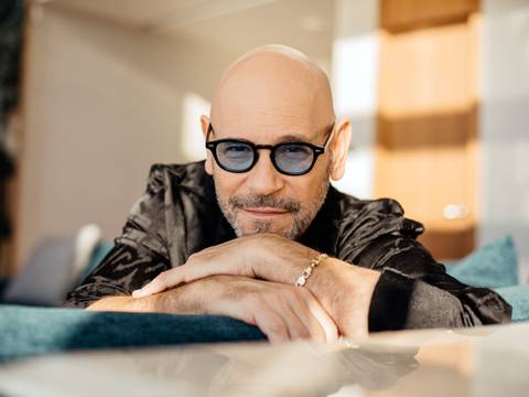 Rami Kleinstein – Männlich gelesene Person mit Glatze und Bart, trägt dunkle Sonnenbrille und schwarze Jacke, stützt sich auf Arme, lächelt leicht, Hintergrund unscharf.