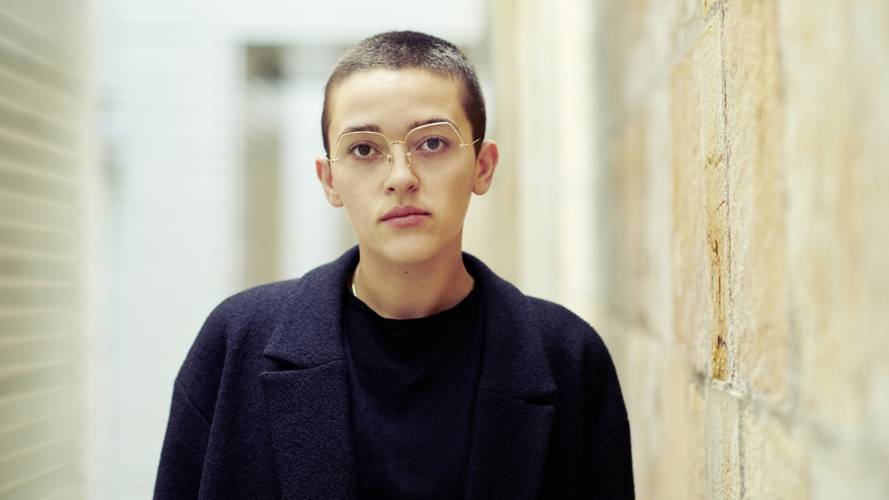 Enis Maci – Person mit Kurzhaarfrisur und Brille, trägt dunklen Mantel, blickt ernst, steht vor einer Stein- und einer Ziegelwand in einem schmalen Gang.