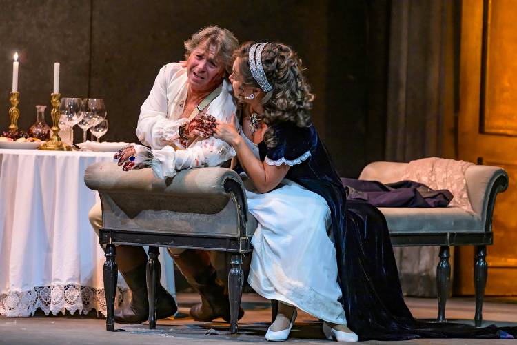 Tosca – Ein sitzender, bekleideter Mann mit blutigen Händen, daneben kniende, bekleidete Frau mit lockigem Haar. Tisch mit Kerzen, Weinflaschen und Gläsern im