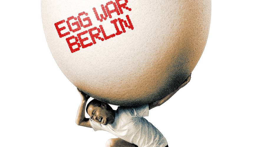 Eine kniende Person trägt ein übergroßes Ei auf den Schultern. Auf der Eierschale steht in roter Schrift „EGG WAR BERLIN“ vor weißem Hintergrund.