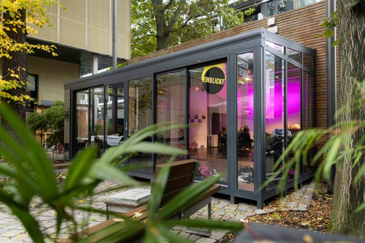 Einblicke Gartenhaus – Moderner Pavillon mit Glaswaenden, innen pink beleuchtet, mit Aufschrift "Einblicke" auf runder Tafel, umgeben von Baeumen und Pflanzen.