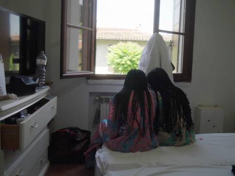 Cem Kaya und Anta Recke, Aus der Sonne, Filmstill © Cem Kaya und Anta Recke – Zwei Personen mit langen Zöpfen sitzen auf einem Bett und schauen aus einem geöffneten Fenster. Draußen sind Häuser und ein Baum sichtbar.