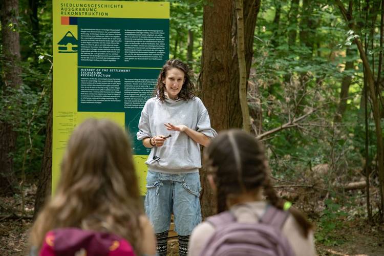 Die Führung informiert unter anderem über die Entstehung des heutigen Museumsdorfs. – Eine Frau erzählt vor einem Schild stehend die Geschichte der mittelalterlichen Siedlung im Museumsdorf Düppel