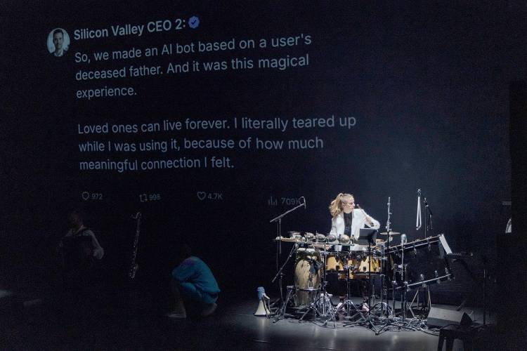 The Alonetimes – Eine Person spielt Schlagzeug vor einer großen Projektion mit dem Text: "Silicon Valley CEO 2: So, we made an AI bot based on a user's deceased father. And it