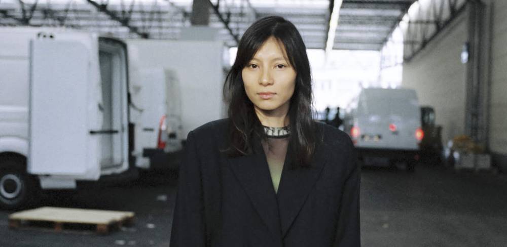 Sung Tieu, Portrait – Portrait einer Person mit langen Haaren in dunkler Kleidung, stehend in einer Halle. Im Hintergrund mehrere geparkte Lieferwagen.