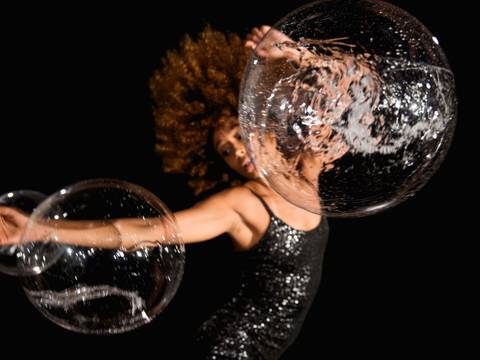 Brazilian Urban Dances – Passinho und Rebolado 1 – Eine Performerin in schwarzen Kleid in Bewegung, vor ihr zwei Seifenblasen.