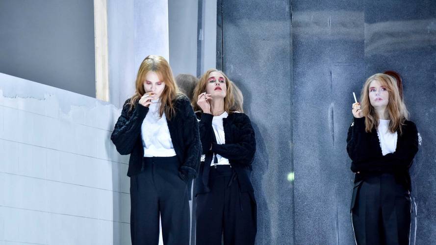 Drei Personen in schwarz-weißen Outfits mit Zigaretten, an Wand gelehnt, mit blonden Haaren und rosa Make-up, in nachdenklicher Pose.