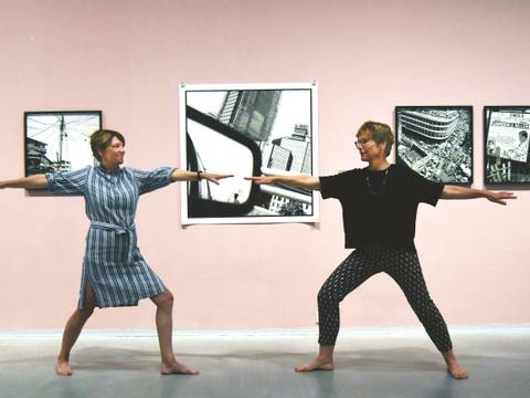 Yoga in der Ausstellung „Akinbode Akinbiyi. Being, Seeing, Wandering“ – Foto: Zwei Personen stehen sich in Yoga-Pose gegenüber und blicken einander an. Im Hintergrund sind schwarz-weiß-Fotografien von Akinbode Akinbiyi vor einer rosafarbenen Wand zu sehen.