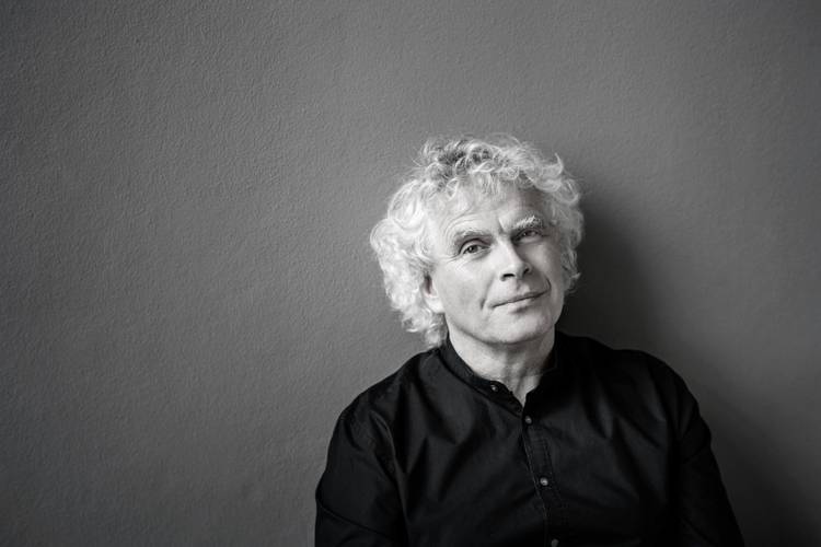 Sir Simon Rattle – Portrait eines Mannes mit grauen lockigen Haaren und schwarzem Hemd vor einem grauen Hintergrund.