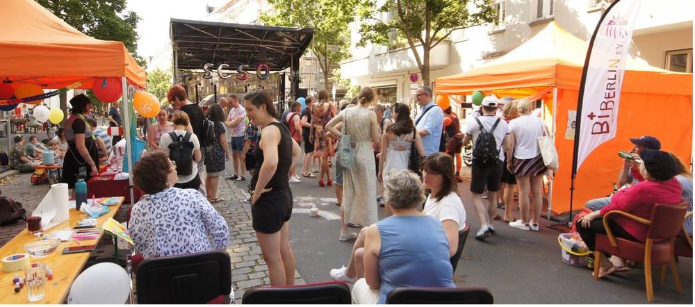 Personengruppe bei Straßenfest mit orangefarbenen Zelten, Luftballons und Informationsstand. Im Hintergrund Bühne mit Schriftzug "SCSO". Rechts Flagge von